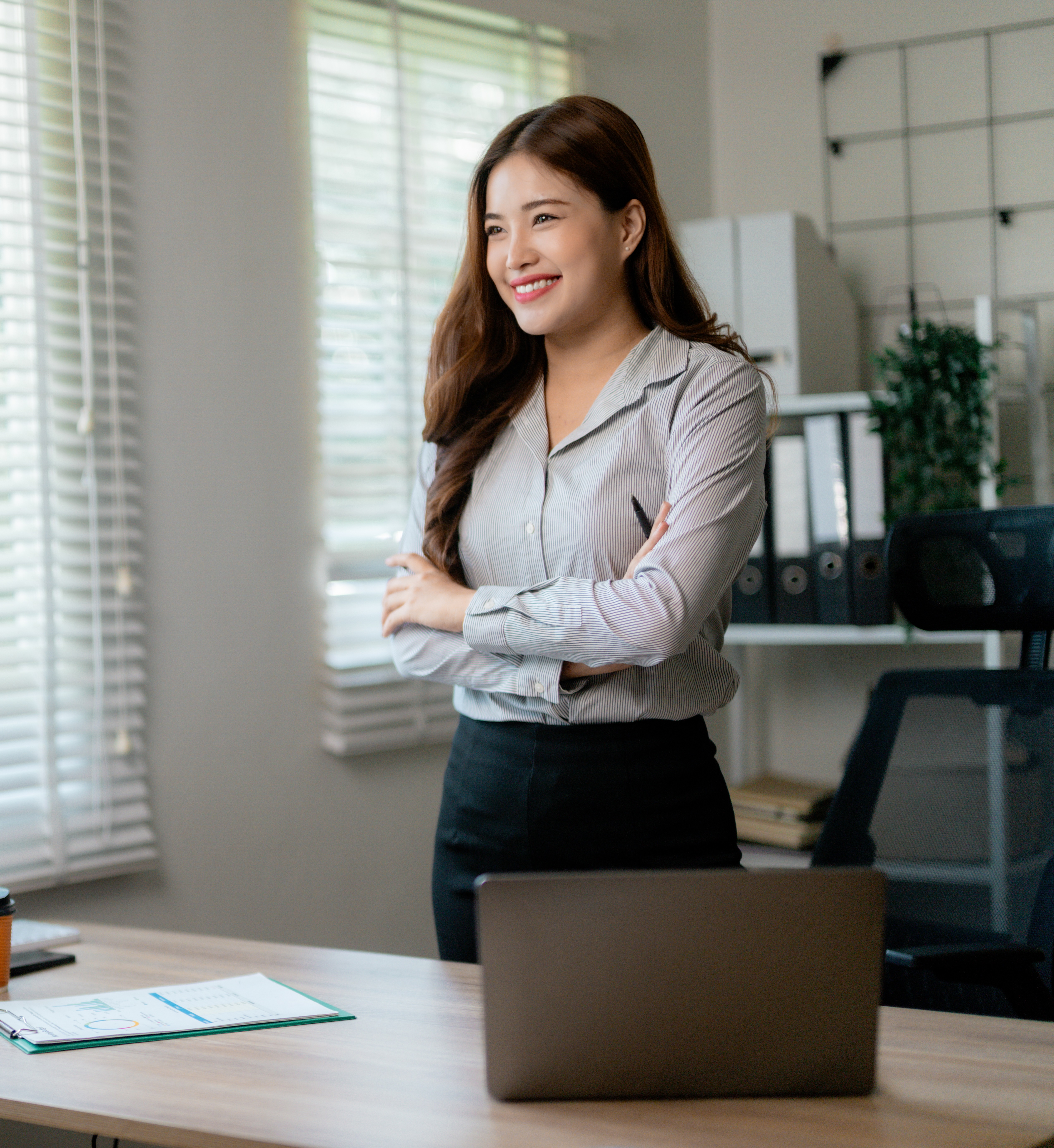 Professional Asian businesswoman smiling confidently, crossing arms near sleek workspace